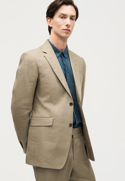 Νεαρός άνδρας με μπεζ κοστούμι και μπλε πουκάμισο με κουμπιά, στεκόμενος μπροστά σε απλό φωτεινό φόντο, κοιτώντας ελαφρώς προς το πλάι.