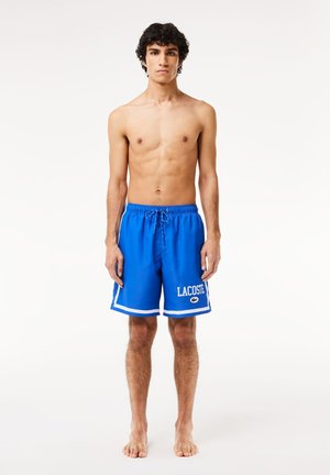 Bañador azul con acentos blancos y cintura con cordón; presenta el logo "LACOSTE" en la pierna izquierda; material ligero y suave.