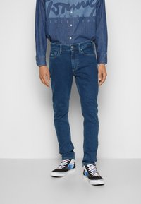 Vaqueros de mezclilla azul con un corte ajustado, que cuentan con cinco bolsillos y detalles de costura sutiles, combinados con zapatillas de deporte con un patrón negro y azul.