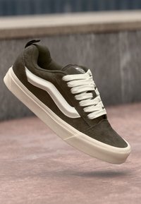 Brown suede sneaker with white accents, classic lace-up design, round toe, and logo on the tongue. Features a textured surface and rubber sole.