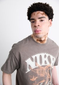 Brun bomulds T-shirt med et stort grafisk print i hvid og orange nuancer. Korte ærmer og en rund hals med et slidt finish.