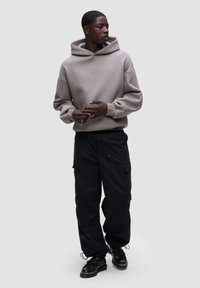 Felpa grigia con cappuccio e spalle scese, abbinata a pantaloni cargo neri larghi con tasche laterali e scarpe nere stringate.