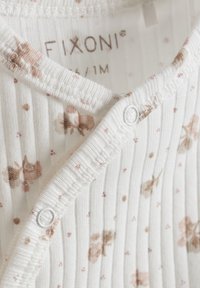 Weißer Baumwoll-Overall mit gerippter Textur, der hellbraune Blumenmuster und pinke Punkte zeigt. Mit weißen Kunststoffsnaps am Ausschnitt befestigt.