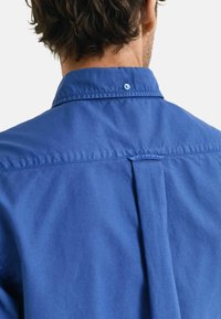 Camicia di cotone blu con colletto button-down, dettaglio con piega sul retro e chiusura a un bottone al collo. Tessuto liscio.