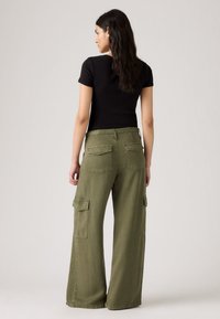 Donna con capelli lunghi e scuri che indossa un top aderente nero a maniche corte e pantaloni cargo verdi oliva larghi, in piedi con le spalle rivolte verso di noi.