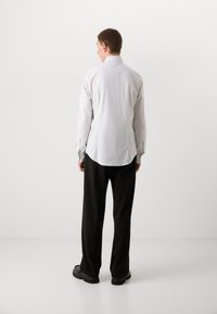 Camisa blanca de manga larga con dobladillo curvado y puños abotonados, combinada con pantalones negros de pierna ancha y zapatos negros. Vista desde atrás.