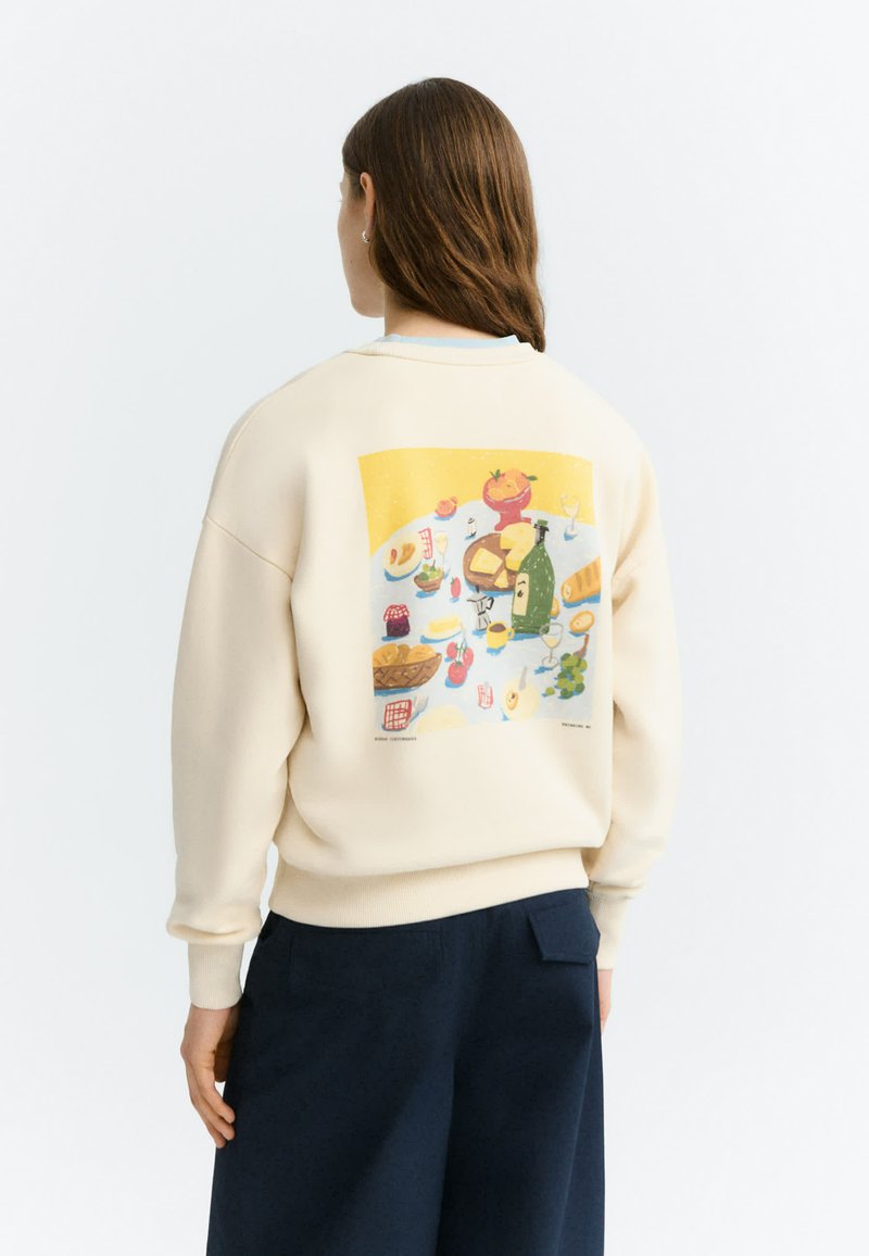 Personne aux cheveux bruns portant un sweat crème avec une illustration colorée d'aliments et de boissons sur une table dans le dos.