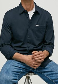 Camicia blu navy con taschino sul petto e etichetta di branding discreta; indossata sopra una maglietta bianca; abbinata a jeans blu.