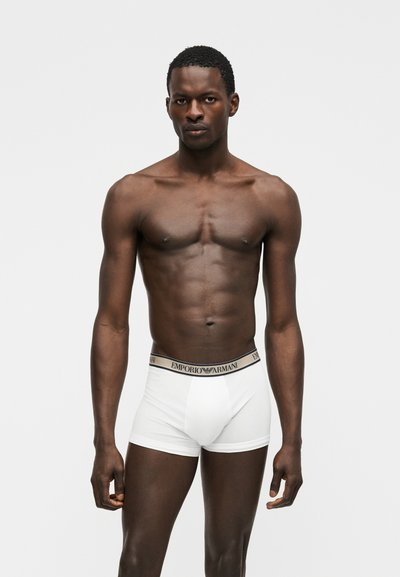 Sötét bőrű férfi modell fehér Emporio Armani boxeralsót viselve, világosszürke egyszínű háttér előtt állva.