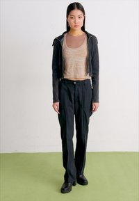 Svart zip-up jacka, beige åtsittande topp och svarta vida byxor. Omrissade händer, kraftiga svarta stövlar, minimalisk design på toppen. Grön golv.