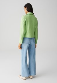 Giacca verde strutturata con colletto, design corto e polsini con bottoni, abbinata a jeans a gamba larga azzurri chiari e scarpe slip-on color crema.