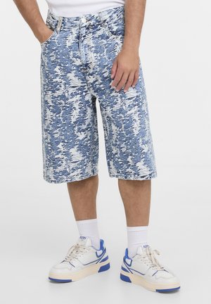 Homme portant un short en denim camouflage bleu et blanc, des baskets blanches avec des accents bleus, et des chaussettes blanches montantes, debout les mains dans les poches.