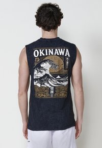 Juodas be rankovių marškinėliai su dideliu baltu ir auksiniu bangų dizainu, tekstu "OKINAWA" ir tekstūruota medžiaga su šoninėmis siūlėmis.