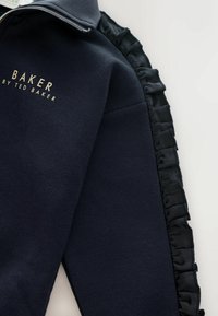 Námořnická mikina s límcem na zip, zlatým logem "BAKER by TED BAKER" a volánkovými saténovými bočními akcenty. Měkká struktura a uvolněný design.