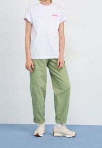 Witte katoenen t-shirt met opgerolde mouwen en roze logo, gecombineerd met lichtgroene cargo-broek en beige sneakers, staande op een blauwe ondergrond.