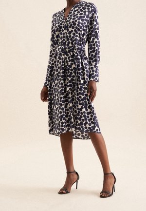 Kvinde iført en knælængde, langærmet kjole med sorte polkaprikker på hvid, kombineret med sorte højhælede sko med ankelrem.