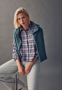 Plaid shirt in purple and blue tones, with a teal fleece vest draped over shoulders; light gray jeans with a smooth texture.