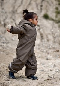 Tuta da neve impermeabile marrone con cerniera e polsini elastici, indossata da un bambino che tiene una pietra mentre cammina su un terreno sabbioso. Stivali di gomma neri.
