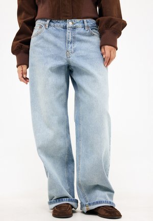 Isik kannab helesiniseid laiade säärega teksapükse, pruune kingi ja pruuni jope, seisab valgel taustal.