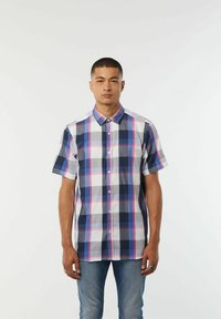 Kortebloesem met een geruit patroon in marineblauw, wit en roze, met een knoopsluiting aan de voorkant en een klassieke kraag. Gecombineerd met een blauwe spijkerbroek.