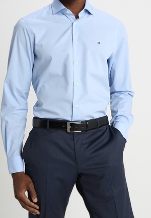 Mężczyzna noszący jasnoniebieską koszulę zapinaną na guziki wpuszczoną w ciemnogranatowe spodnie z czarnym skórzanym paskiem, jedna ręka w kieszeni.