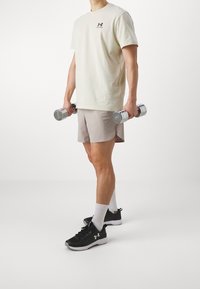 Ljusbeige t-shirt med logotyp, ljusgrå shorts, svarta sneakers med vita accenter, håller silvriga hantlar, står på en neutral bakgrund.