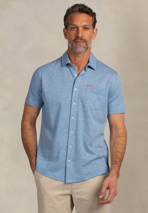 Man met grijze baard, gekleed in een lichtblauw overhemd met korte mouwen en beige broek, staande met één hand in de broekzak tegen een beige achtergrond.