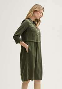Olivengrünes Kleid mit Knopfleiste, Brusttaschen und Dreiviertelärmeln. Hergestellt aus einem glatten Stoff, bietet es eine lässige Silhouette.