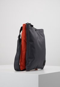 Šedá a oranžová taška přes rameno vyrobená z odolné tkaniny, s uzávěrem na zip, nastavitelným popruhem a minimalistickým designem s logem.