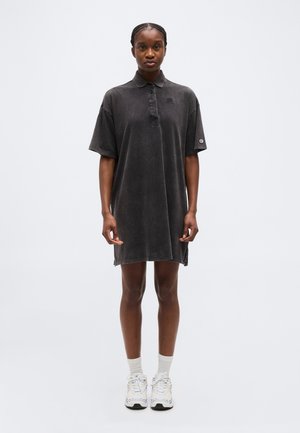 Kobieta stoi, ubrana w luźną czarną sukienkę polo i białe trampki z skarpetkami, na tle jasnego, jednolitego tła.