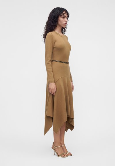 Une femme porte une robe en maille brun camel à manches longues et à l’ourlet asymétrique, avec une ceinture chaîne et des sandales dorées à talons hauts, sur un fond uni.