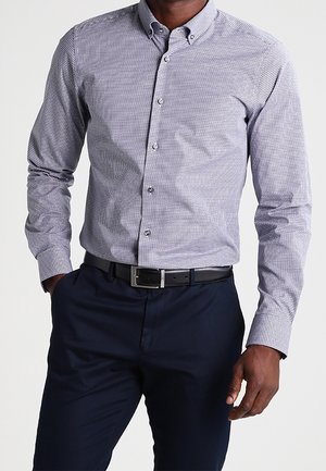 Homme portant une chemise boutonnée gris clair à motifs rentrée dans un pantalon bleu marine foncé avec une ceinture en cuir noire.