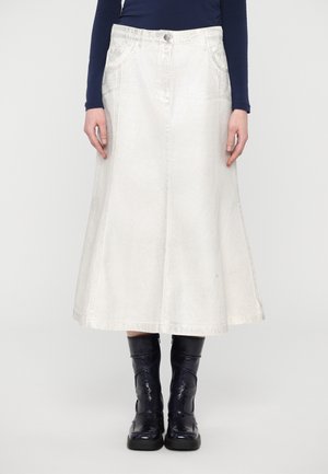Femme portant une jupe blanche mi-longue avec des poches sur le devant, un haut bleu marine à manches longues et des bottines noires brillantes.