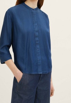 Žena nosící volnou modrou denimovou blůzu s tříčtvrtečními rukávy a tmavě modré denimové kalhoty, jednou rukou v kapse.