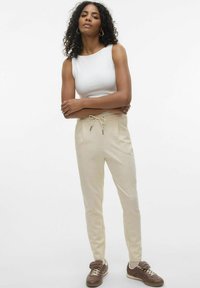 Lichtbeige joggers met een trekkoord in de taille, zijzakken en een taps toelopend ontwerp, gecombineerd met een witte mouwloze top en bruine sneakers.