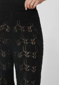 Zwarte gebreide broek met een ingewikkeld opengewerkt kantontwerp, met golvende patronen en een geribde tailleband. Detailopname benadrukt de textuur.