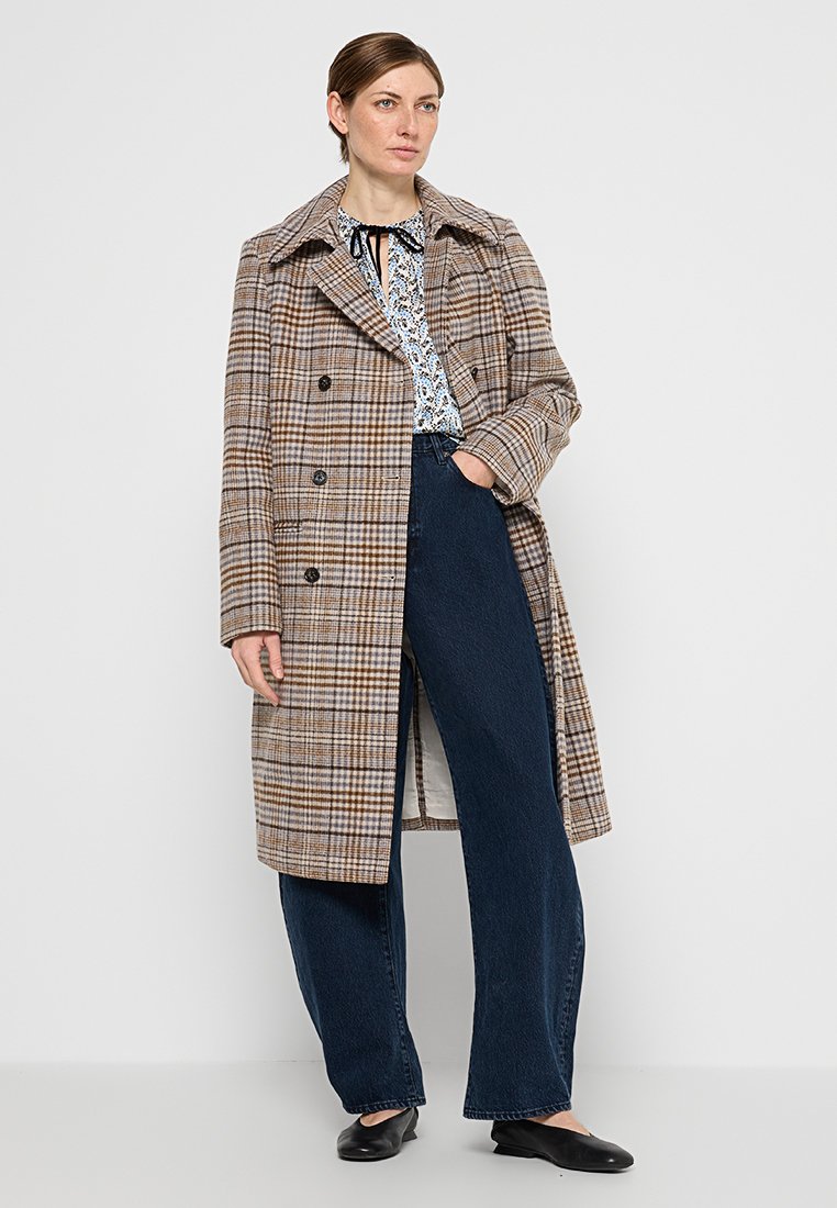 Bruine geblokte overjas met een dubbelzijdig ontwerp, grote revers en zijzakken, gedragen over een blauwe bloemenblouse en donkere spijkerbroeken.