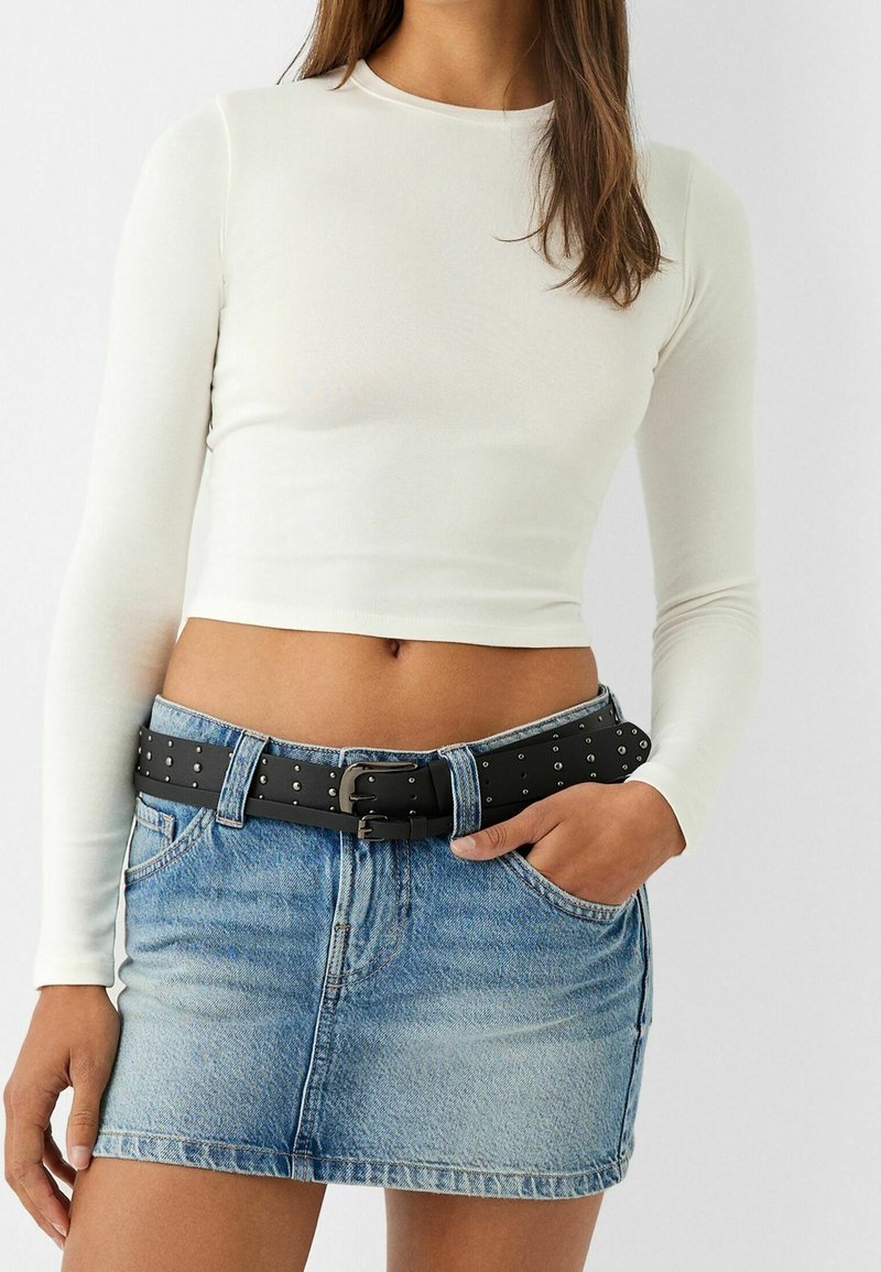 Kvinna i en vit långärmad cropped topp, ljusblå denim minikjol och svart nitarbälte med dubbla spännen, hand i fickan.
