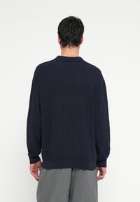 Laivastonsininen neulepaita, jossa pystysuora ribbineulos, kauluksellinen pääntie, väljä istuvuus ja leveät hihat, yhdistetty harmaisiin housuihin.