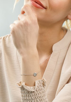 Silver bracelet with a circular charm on a delicate chain, worn on a light-skinned wrist, paired with a light, textured blouse.