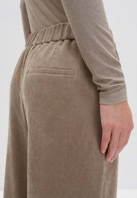 Gros plan sur une personne portant un pantalon beige côtelé avec une taille élastique et une poche arrière passepoilée, se tenant avec le bras détendu sur le côté.
