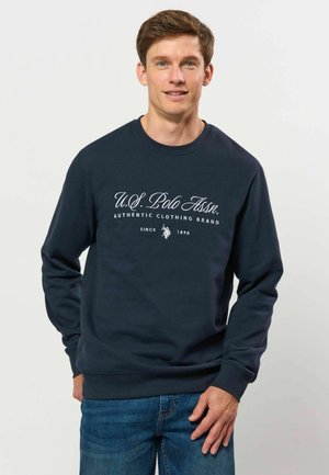 Homme portant un sweat-shirt bleu marine avec le texte "U.S. Polo Assn. Authentic Clothing Brand" et un jean bleu devant un fond clair uni.