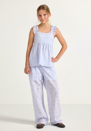 Nuori tyttö seisoo käsi lanteilla, yllään vaaleansininen, silmukkakuvioinen hihaton paita, jossa on röyhelöiset olkahihnat, ja siihen sopivat leveälahkeiset housut.