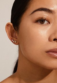 Tre orecchini in un orecchio: due perle bianche e uno perno d'oro con una superficie texture, su uno sfondo di pelle liscia.