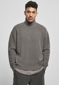 Jersey de cuello alto gris con un tejido texturizado, corte holgado y puños y dobladillo acanalados, superpuesto a una camisa gris clara.