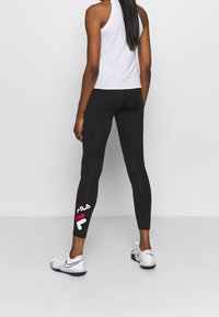 Schwarze Leggings mit glatter Textur, mit einem weißen und roten FILA-Logo an der unteren linken Beinseite. Kombiniert mit einem weißen ärmellosen Oberteil und weißen Sneakers.