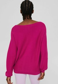 Personne aux cheveux tressés portant un pull ample rose vif et un pantalon rose clair, vue de dos sur un fond uni.