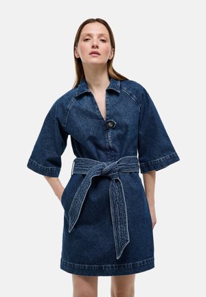 Femme portant une robe en jean bleu foncé avec des manches larges, des boutons sur le devant et une large ceinture nouée à la taille.