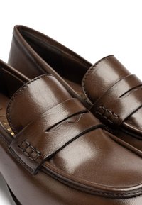 Mocasines de cuero marrón con un diseño elegante, puntera redondeada, acentos de costura elevados y una textura sutil en la superficie.