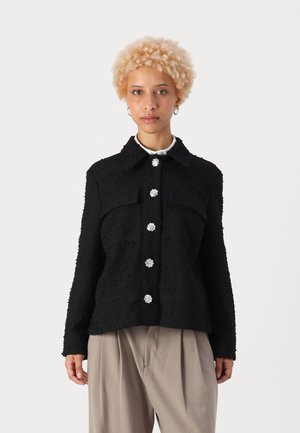 Black textured jacket with a collar, featuring silver flower-shaped buttons and two chest pockets. Worn over beige trousers.